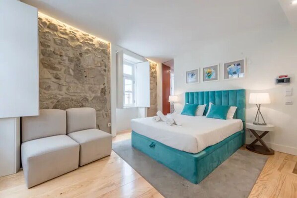 Un dormitorio moderno con una pared de acento de piedra, una cama grande con ropa de cama color turquesa, un sofá blanco y una mesita de noche pequeña.