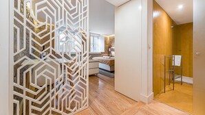 A modern hotel room with a geometric white partition, a wooden floor, a bed, a bathroom with a wooden door, and a shower area.