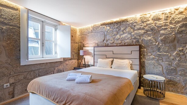 A bedroom with a stone wall, a bed with a headboard, a bedside table, and a window with blinds.