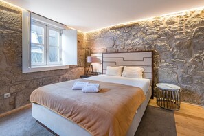 Un dormitorio con pared de piedra, una cama con cabecera, una mesita de noche y una ventana con persianas.