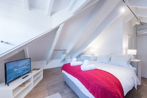 Una habitación de hotel moderna con techo inclinado, una cama con ropa de cama blanca y roja, un televisor sobre un soporte blanco y piso de madera.