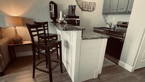 A compact kitchen with a central island, dark wooden chairs, a microwave, a kettle, and a sink. - Chill Beach front complex Two Queens at Top of the Gulf (Panama City Beach)