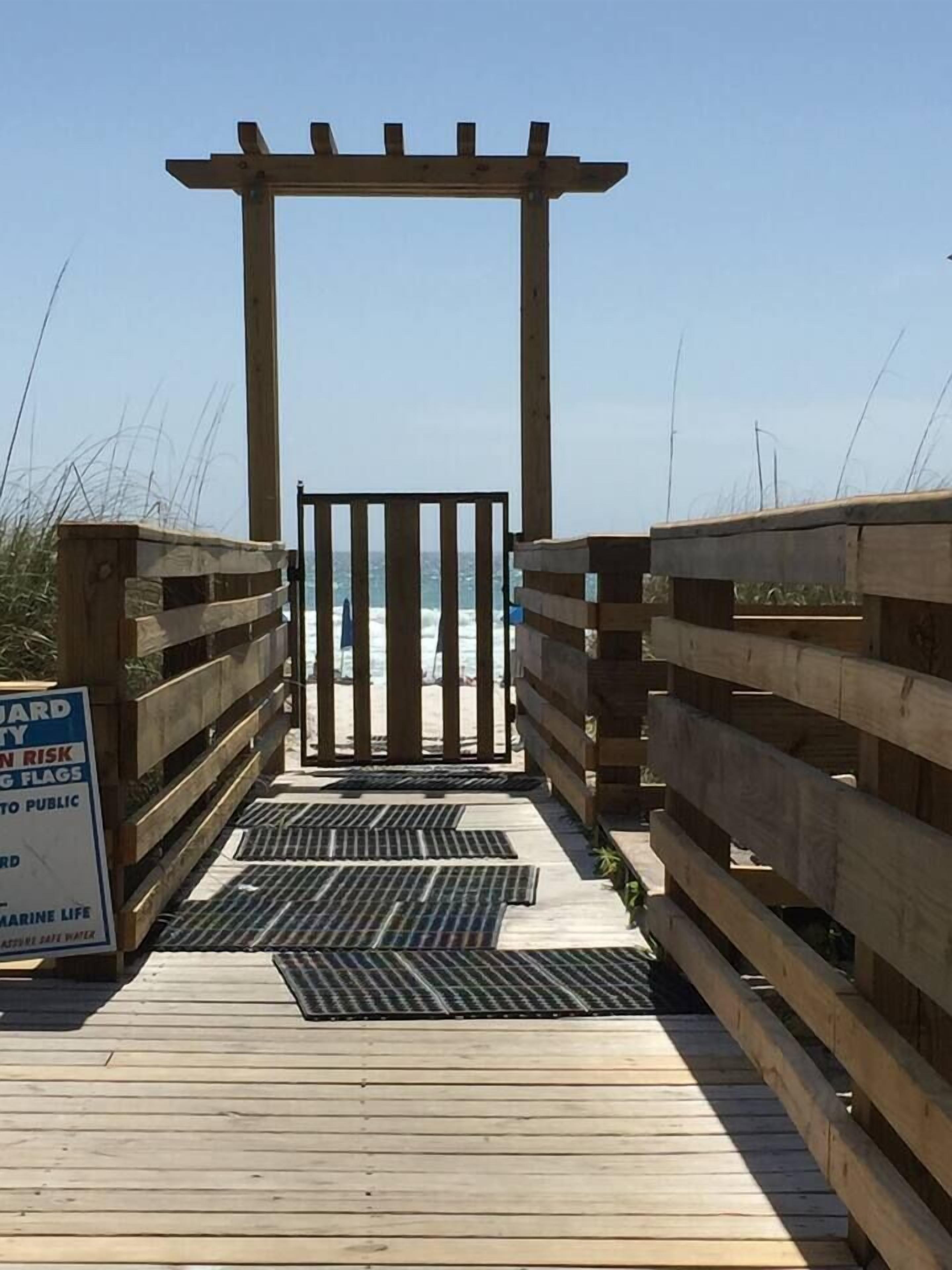 Ein hölzerner Steg führt zu einem Strand, mit einem Schild, das vor Meereslebewesen und Gezeitenrisiken warnt.