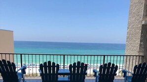 Un balcón con tres sillas Adirondack y una mesa, con vista a una playa y una visión despejada del océano.