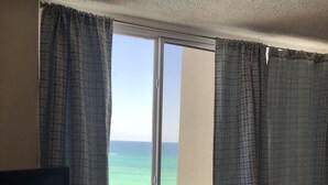 Una habitación con un ventanal que da a la playa, un televisor de pantalla plana sobre un soporte de madera, y una cama con colcha estampada.