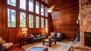 A cozy log cabin interior with a stone fireplace, wooden floors, a leather sofa, and large windows offering a view of the forest.