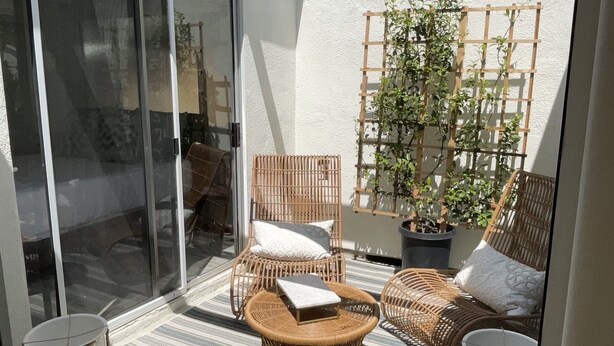 Een zonnig terras met rieten meubilair, een bloempot en een glazen schuindeur naar een binnenruimte.