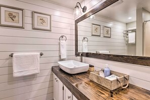 Une salle de bain avec un lavabo blanc, un plan de toilette en bois et un grand miroir. Des tableaux encadrés ornent le mur, et il y a aussi un porte-serviettes.