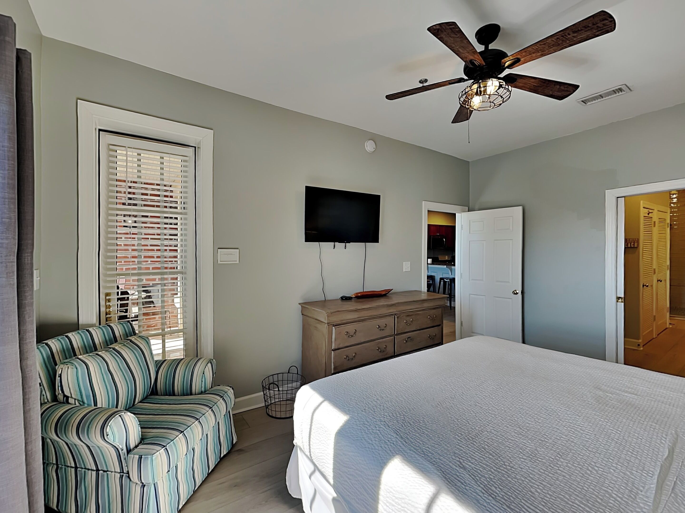 A bedroom with a bed, a TV, a ceiling fan, a dresser, and a window with blinds.