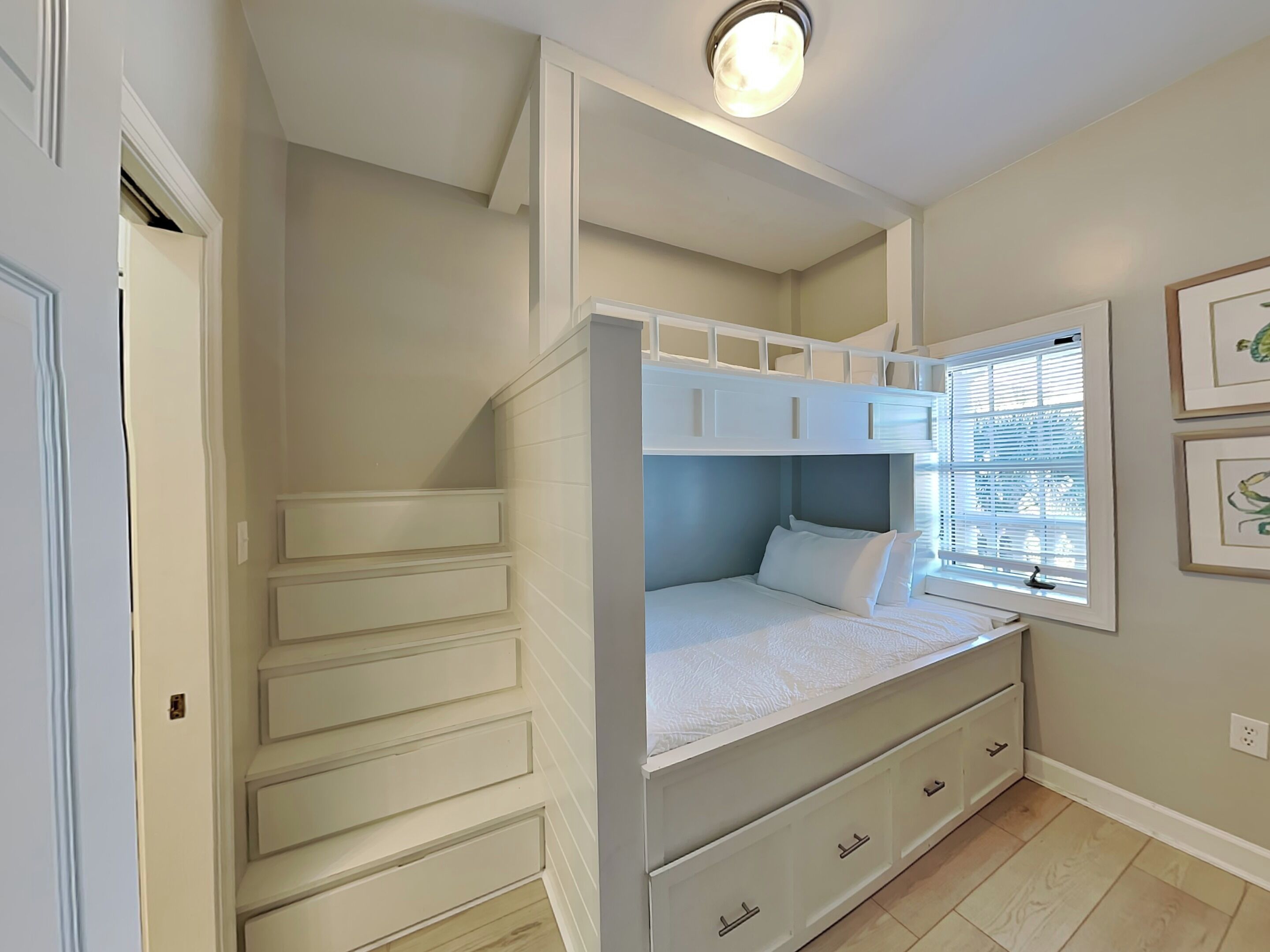 A bedroom with a bunk bed, built-in drawers, a window with blinds, and a light fixture.