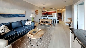 A modern living room with a blue sofa, a wooden coffee table, and a dining area with a kitchen in the background. - Village Of South Walton #263A | Luxe Condo in the Village! (Seacrest Beach)
