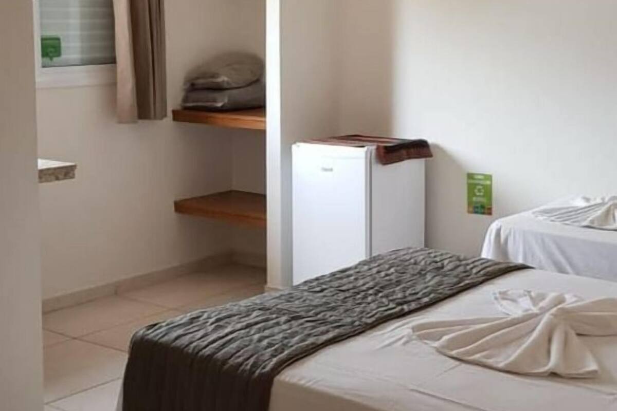 Un dormitorio pequeño y ordenado, con una cama, un refrigerador compacto y un estante con ropa doblada.