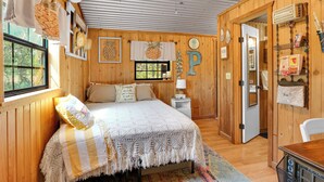 Una habitació de fusta acollidora amb un llit, una tauleta de nit, una làmpada i una porta que dona a una altra habitació.