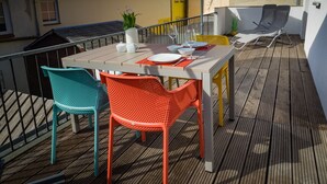 Un espace de restauration en plein air avec une table dressée pour deux personnes, entourée de chaises colorées, d’une petite plante en pot et d’un parasol.