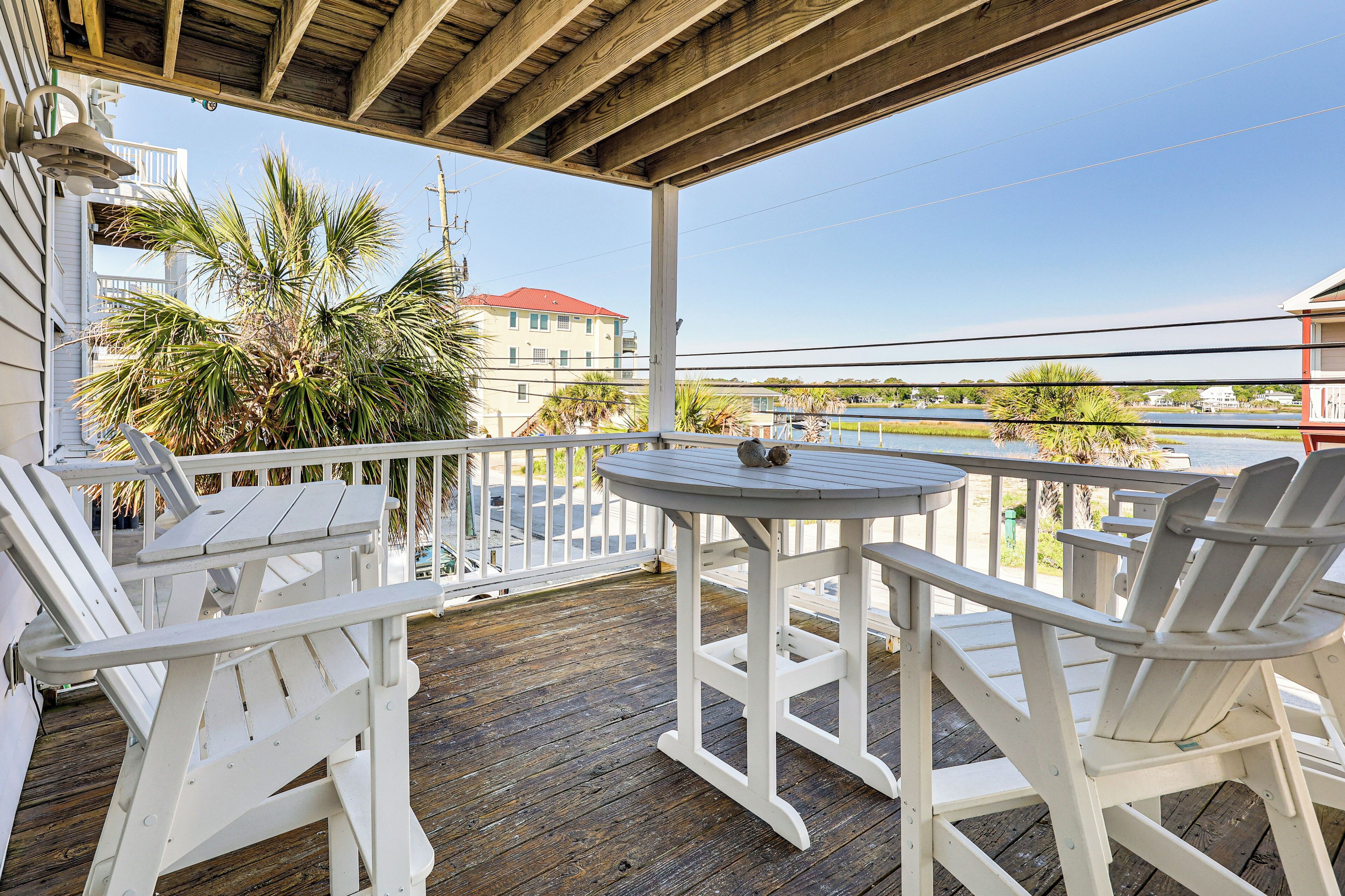 Une terrasse en bois avec des chaises Adirondack blanches et une petite table, d’où l’on peut admirer un plan d’eau et des bâtiments.