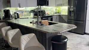 Eine moderne Küche mit weißen Schränken, einem Edelstahlkühlschrank, einer Mikrowelle, einer Kaffeemaschine und einem Spülbecken. Es gibt drei weiße Barhocker und einen Mülleimer.