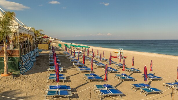 A beach with rows of blue lounge chairs and red umbrellas, a sandy shore, and a clear blue sky. - La Casa Dei Ricordi Letojanni (Letojanni)