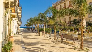 A paved street lined with palm trees and benches, leading to a building with balconies and outdoor seating. - La Casa Dei Ricordi Letojanni (Letojanni)