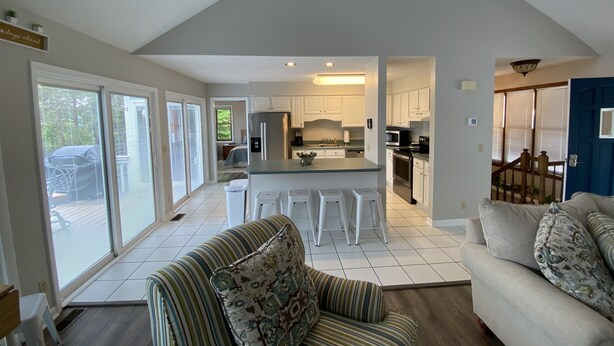 A spacious living area with a large window, a kitchen with white cabinets, and a dining area with bar stools. - Little Buck Bungalow - Deerfield Lake View Home on Norris Lake (LaFollette)