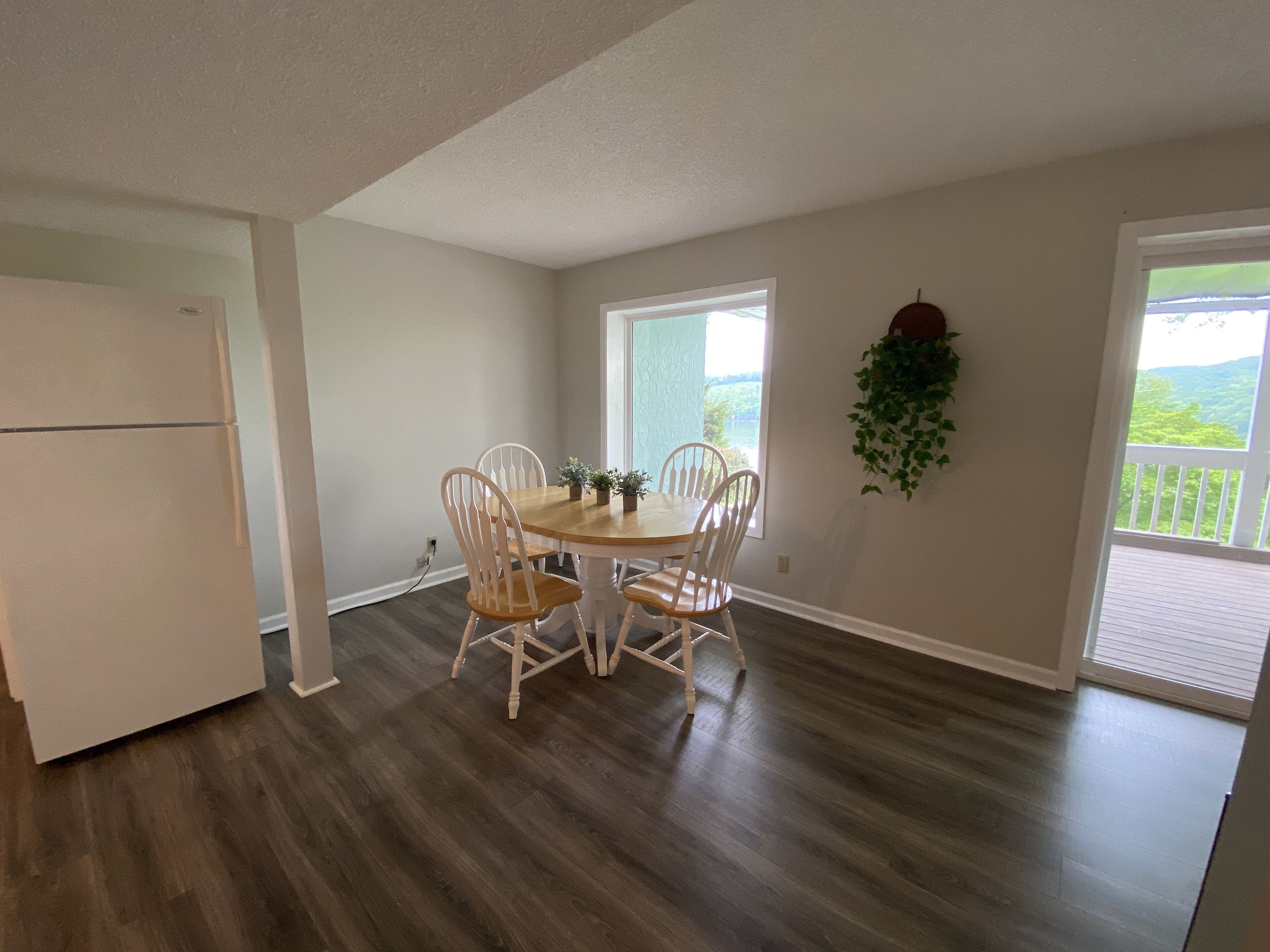 A dining area with a round table and four chairs, a white refrigerator, and a view of greenery through large windows.