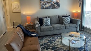 Ein Wohnzimmer mit grauem Sofa, einem braunen Ledersessel, einem Glastisch und einem gemusterten Teppich. An der Wand hängt ein Gemälde und eine Tür führt in einen anderen Raum.