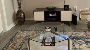 Ein Wohnzimmer mit einem Glastisch, einem an der Wand befestigten Flachbildfernseher, einer dekorativen Vase, einer Holzkommode und einem gemusterten Teppich.