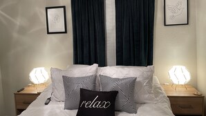 Ein ordentlich bezogenes Bett mit einem schwarzen, mit „relax“ beschrifteten Kissen und gemusterten Kissen, flankiert von Nachttischen mit Lampen.