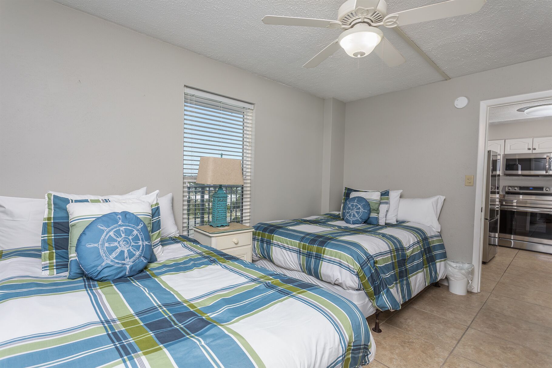A bedroom with two beds, a ceiling fan, a window with blinds, and a kitchenette area.