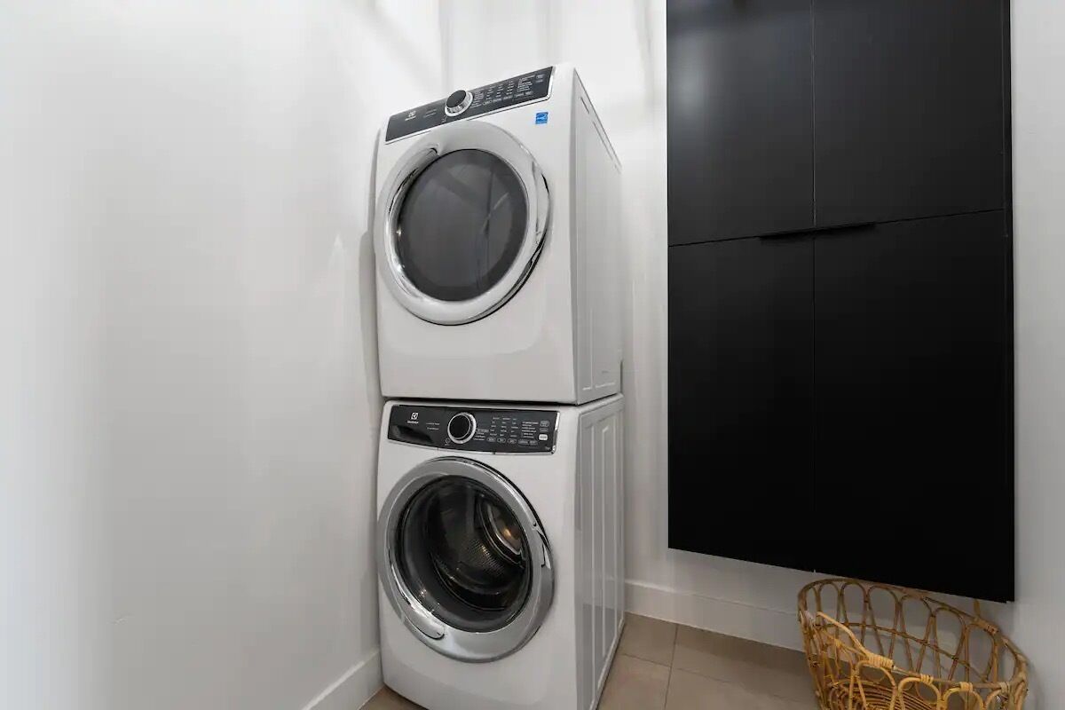 Un appareil combiné lave-linge et sèche-linge, placé dans un coin de la pièce, avec un meuble noir et un panier en osier.