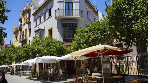 A row of outdoor cafe tables and chairs with umbrellas, set against a backdrop of a white building with balconies.