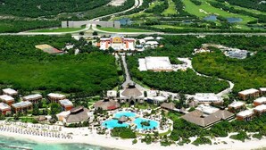 Vista aérea de un complejo turístico con varias edificaciones, una gran piscina central y una zona de playa.