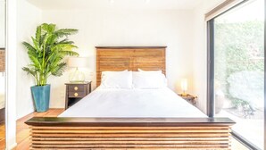 Un dormitorio moderno con una cama grande, una planta en maceta, una mesita de noche con lámpara y vistas al verdor exterior.