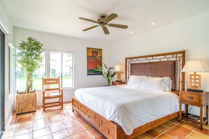 A bedroom with a large bed, a ceiling fan, a wooden nightstand, a chair, and a potted plant.