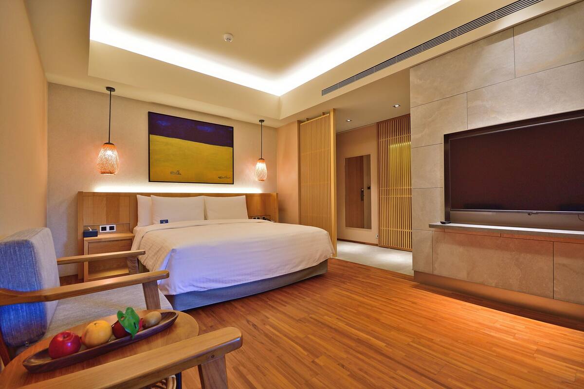 A modern hotel room with a large bed, wooden flooring, a flat-screen TV, and a fruit bowl on a wooden side table.