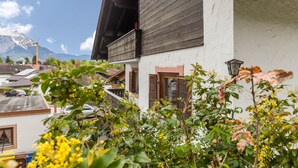 Edifici tradicional amb un balcó, una muntanya al fons i un jardí al primer pla.