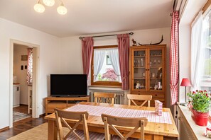 Une pièce comprenant une table à manger en bois, des chaises, un téléviseur, un meuble avec des portes en verre et une fenêtre avec des rideaux.