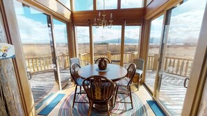 Un coin repas avec une table ronde et des chaises, de grandes fenêtres offrant une vue sur un paysage montagneux, et une terrasse en bois à l’extérieur.