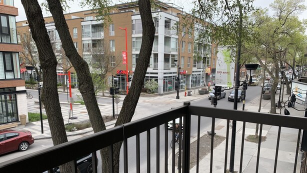 Vista desde un balcón sobre una calle con árboles, un coche rojo y un edificio con un cartel que dice 'Vivere 100 anni'.