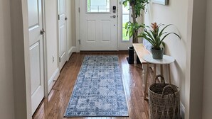 Un pasillo con piso de madera, un tapete azul con estampado, una puerta blanca y una pequeña mesa con una planta en maceta.