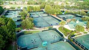 Vista aérea de un complejo deportivo con varias canchas de tenis rodeado de árboles y un campo de golf al fondo.