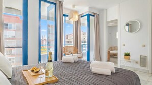 A hotel room with a bed, a tray with a bottle and glasses, a chair, and a view of buildings through large windows. - Marbella Pool House PH78 On Sea Shore, Marbella, Spain (Marbella)