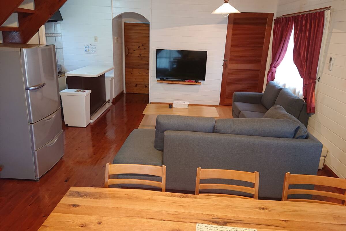 Ein kompakter Wohnbereich mit Esstisch aus Holz, grauem Sofa und einem an einer weißen Wand montierten Fernseher.
