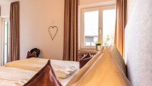 Hotellihuone, jossa on sänky, sydämenmuotoinen koriste seinällä, ikkuna, josta avautuu näkymä vehreään maisemaan, ja ruukkukasvi ikkunalaudalla.