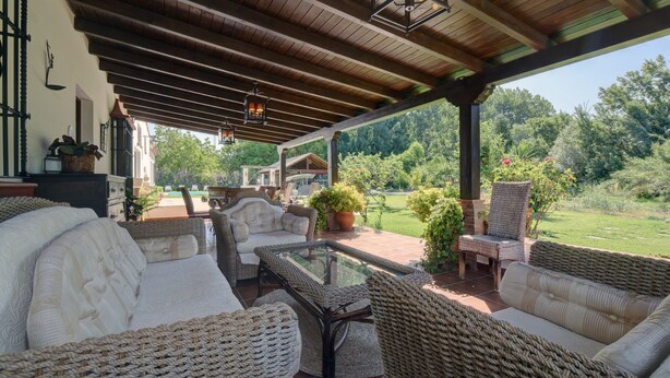 A covered outdoor seating area with wicker furniture, a glass-top table, and a view of a lush garden.