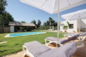 Uma área junto à piscina com espreguiçadeiras e guarda-sóis, uma piscina e um alpendre coberto.