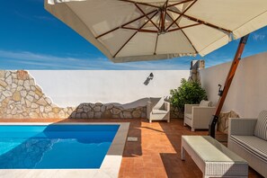Une piscine bordée d’un muret en pierre, dotée de meubles en osier et d’un grand parasol.