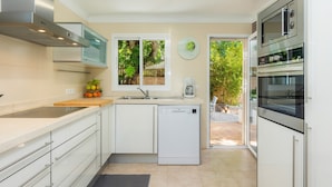 A modern kitchen with white cabinetry, a dishwasher, a double oven, and a window overlooking a garden.