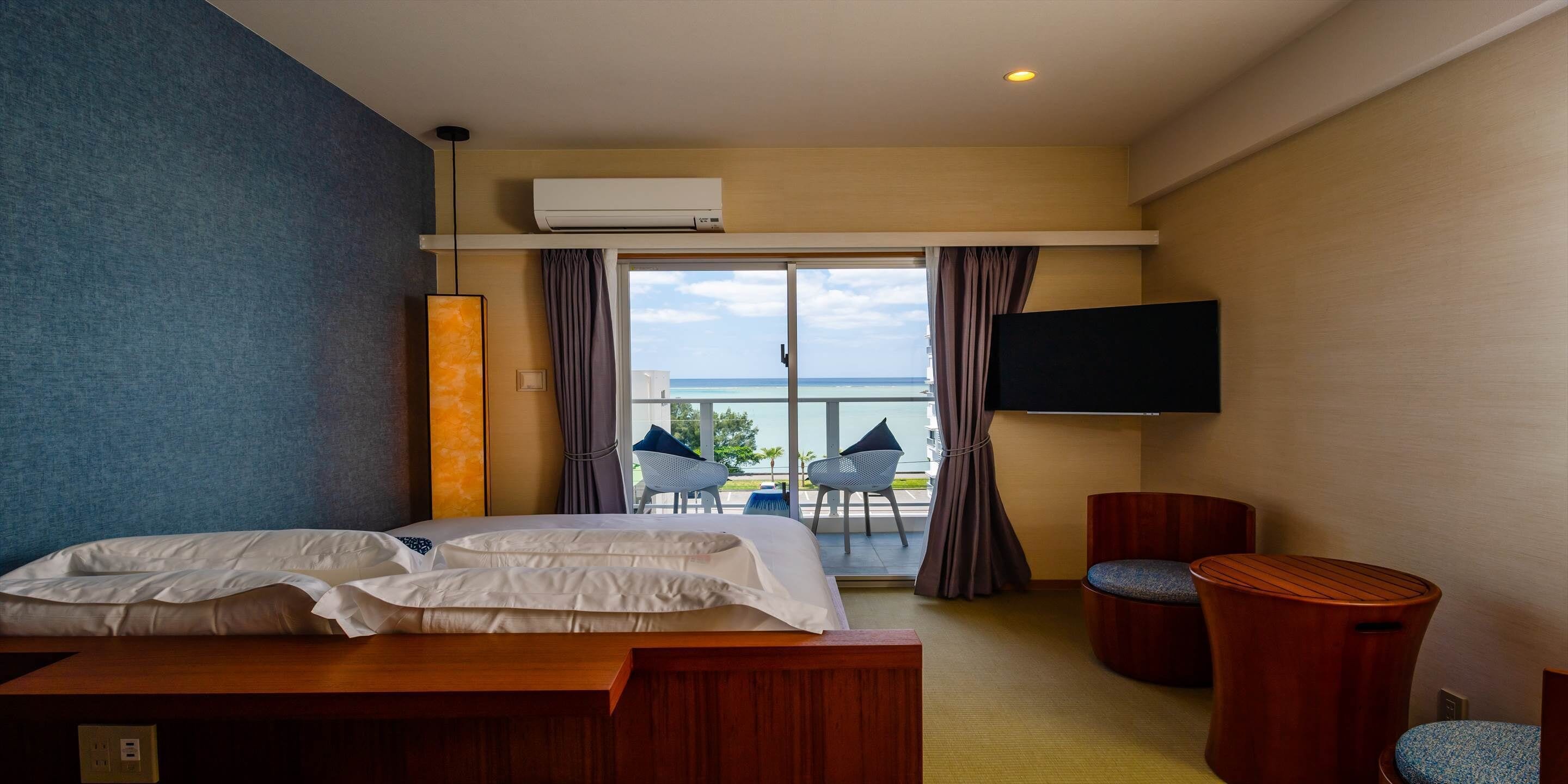 A hotel room with a bed, a TV, a balcony with chairs, and an air conditioning unit.