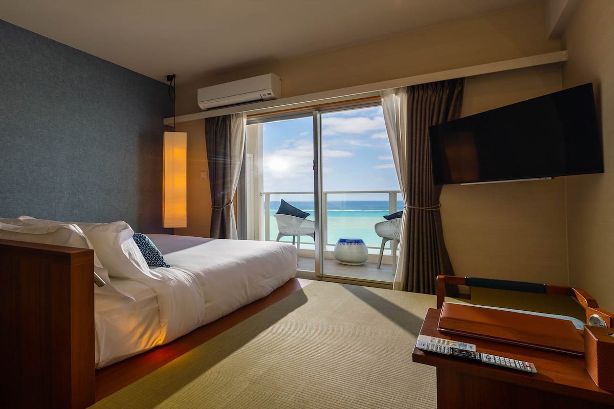 A hotel room with a bed, a desk, a chair, a TV, and a balcony with a view of the sea.