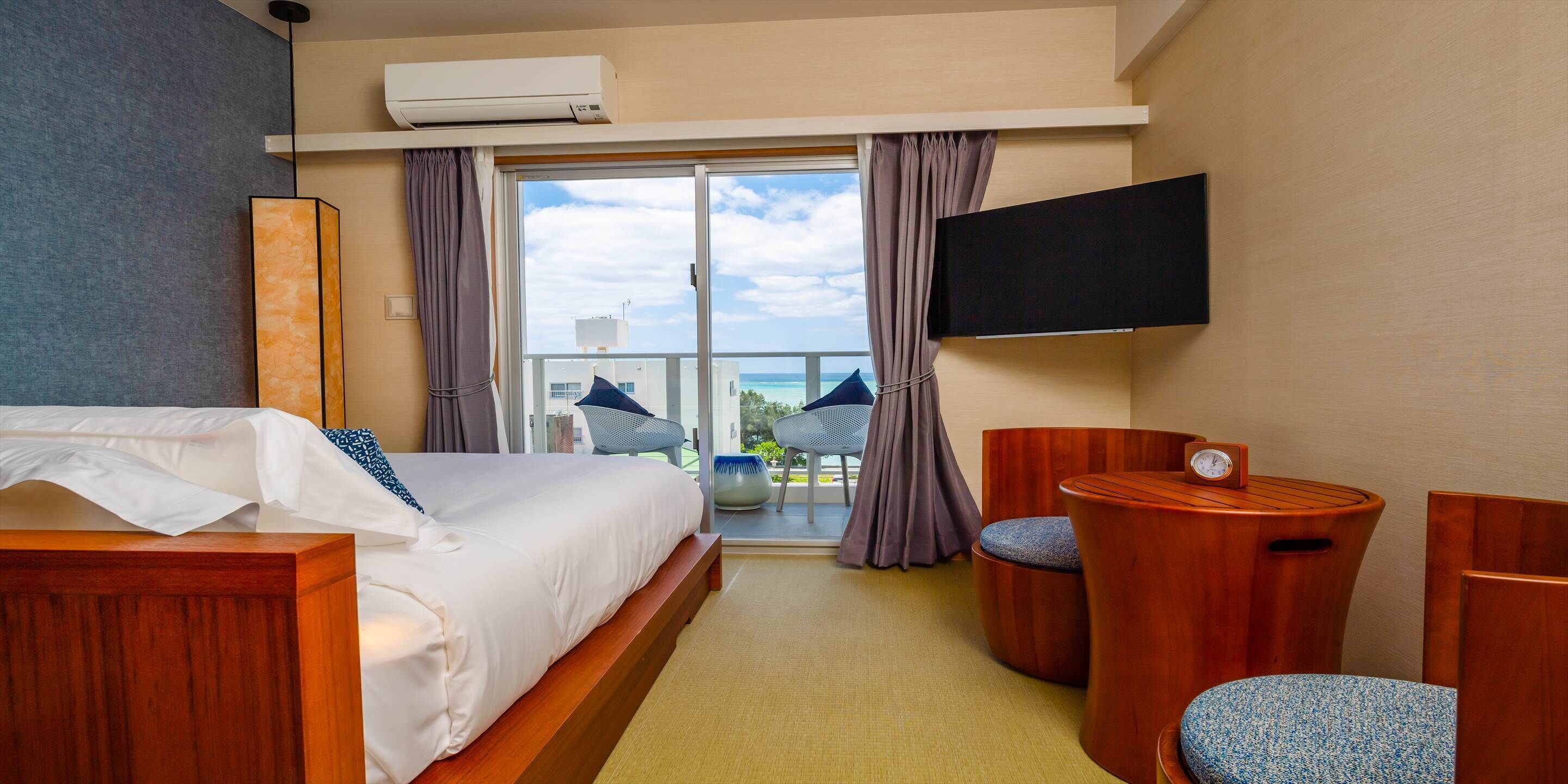 A hotel room with a bed, a TV, a small table with a clock, and a balcony with a view.